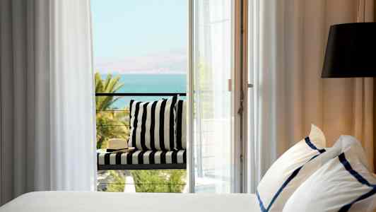 Seng med hvide og blå lagner ved siden af en altan med udsigt over havet. (AI-genereret tekst)