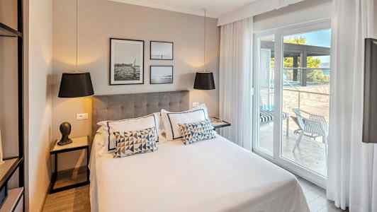 Moderne hotelværelse med dobbeltseng, dekoreret med puder og kunst på væggen. Glasdøre fører ud til en møbleret terrasse. (AI-genereret tekst)