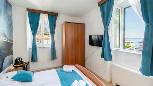 Hotelværelse med dobbeltseng, blå gardiner og udsigt til en havn udenfor vinduet. (AI-genereret tekst)