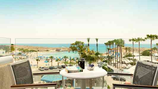 Udsigt fra en altan med bord og stole mod en strand med parasoller og palmer ved havet. (AI-genereret tekst)