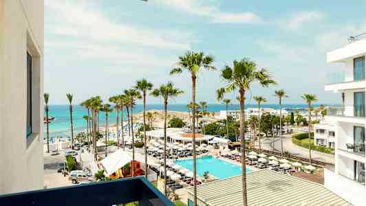 Udsigt over en strand med palmer, en pool og en hvid bygning ved havet. (AI-genereret tekst)