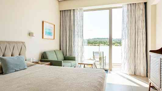 Et hotelværelse med en seng, sofa og bord samt en balkon med stole og udsigt over grønne områder. (AI-genereret tekst)