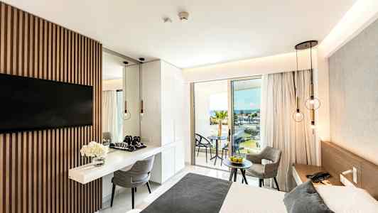 Moderne hotelværelse med en dobbeltseng, bord og stole samt udsigt over havet fra en balkon. (AI-genereret tekst)