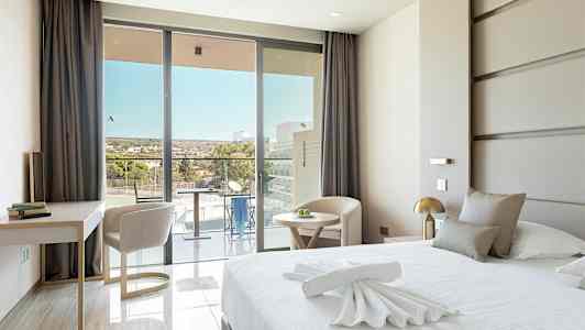 Et moderne hotelværelse med en stor seng, et skrivebord og stole samt en balkon med udsigt. (AI-genereret tekst)