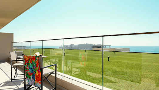 En terrasse med glasrækværk, stole og udsigt over grønne græsplæner og havet. (AI-genereret tekst)