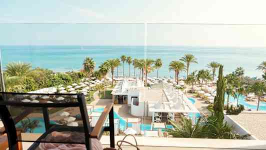 Udsigt over havet, hotelområde med palmer og swimmingpools, og en stol i forgrunden. (AI-genereret tekst)