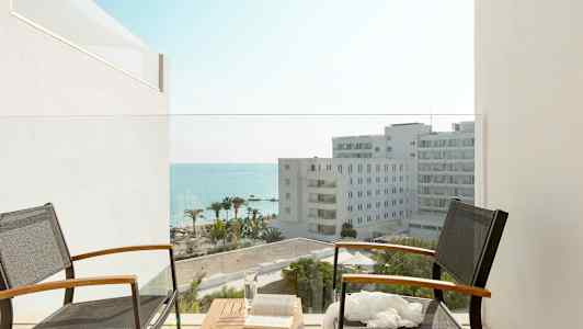 To stole med bord på altan med udsigt til hotelbygninger, palmer og hav. (AI-genereret tekst)