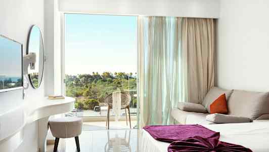 Et lyst hotelværelse med en sofa, en seng med lilla tæppe og en altan med udsigt over grønt område. (AI-genereret tekst)