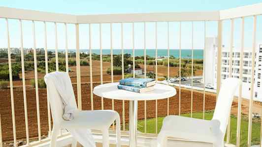 To hvide stole og et bord med bøger på en altan med udsigt over landbrugsjord og havet. (AI-genereret tekst)