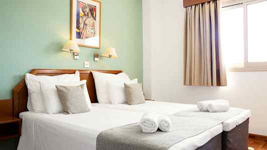 Interiør af et hotelværelse med dobbeltseng, to sengelamper, et vindue med gardin og et dekorativt vægmaleri. (AI-genereret tekst)