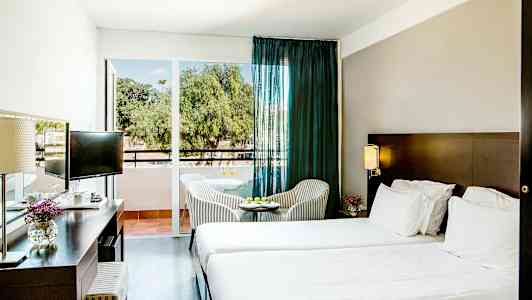 Hotelværelse med to senge, et skrivebord og et lille bord med stole foran en glasdør. (AI-genereret tekst)