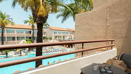 Udsigt fra en altan med bord og stole over en pool og omkringliggende bygninger. Palmer ses i baggrunden. (AI-genereret tekst)