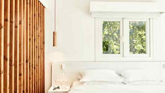 Lyst og luftigt soveværelse med en seng, hvidt sengetøj og en træpanel samt vinduer med udsigt til grønne omgivelser. (AI-genereret tekst)