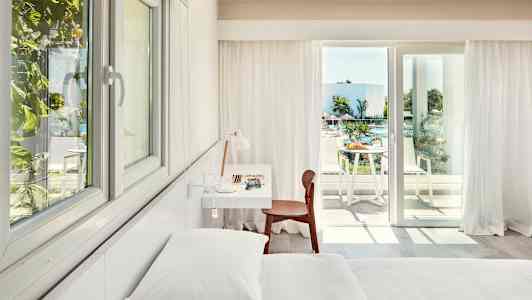 Interiør af et lyst hotelværelse med seng, skrivebord og stol samt en balkon med udsigt over en pool og grønne områder. (AI-genereret tekst)