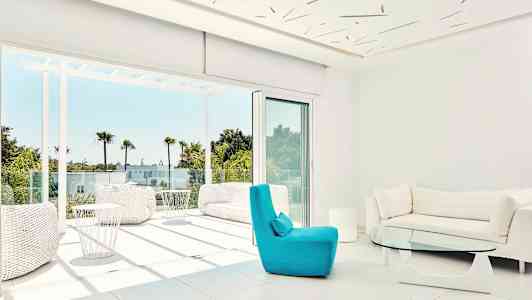 Moderne stue med store vinduer og udsigt til en solrig terrasse med palmer. (AI-genereret tekst)