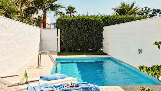 En mindre pool omgivet af hvide vægge og grøn vegetation, med to solsenge nær kanten. (AI-genereret tekst)