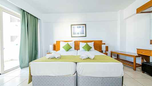 To enkeltsenge med grønt sengetæppe og hvide puder i et hotelværelse med lys indretning. (AI-genereret tekst)