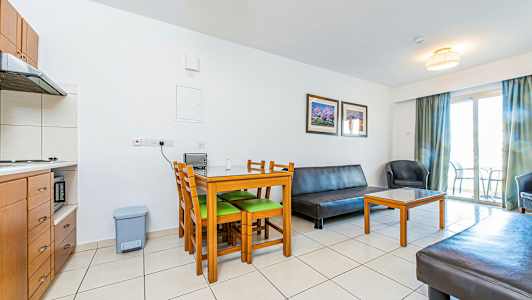 En stue med møbler inklusive et bord med stole, en sofa og en lænestol. Et køkken kan ses i hjørnet og en altan med dør i baggrunden. (AI-genereret tekst)