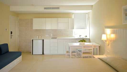 En lys og moderne studio med køkkenområde med hvide skabe, et lille bord med stole og en siddemøbel mod en beige væg. (AI-genereret tekst)