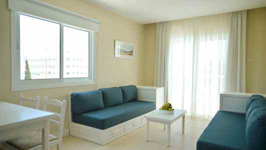 Et værelse med to sofaer med blå puder, et hvidt bord og et stort vindue med hvide gardiner. (AI-genereret tekst)