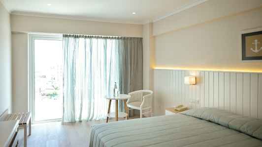 Billede af et lyst hotelværelse med en seng, et bord og en stol nær et vindue med lyse gardiner. (AI-genereret tekst)