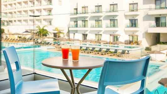 To farverige drikkevarer står på et bord ved siden af to stole, med en pool og et hotel i baggrunden. (AI-genereret tekst)