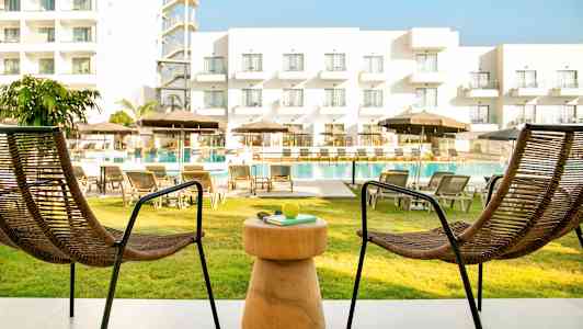 To stole og et bord på en terrasse med udsigt over poolområdet og et hotel. (AI-genereret tekst)