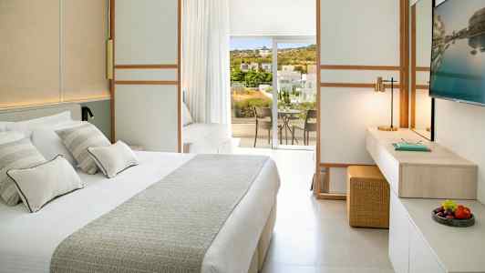 Et lyst hotelværelse med en stor seng, et arbejdsområde og en balkon med udsigt over grønne områder og bygninger. (AI-genereret tekst)