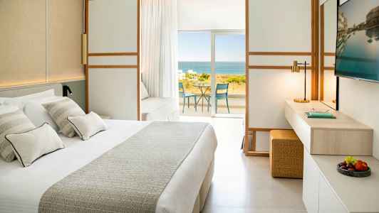 Et hotelværelse med en stor seng, TV og udsigt over havet fra en balkon. (AI-genereret tekst)