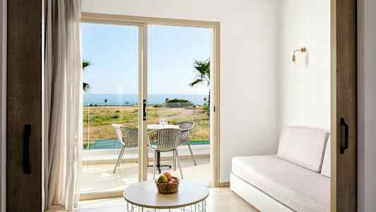 Et rum med sofa, bord og glasdøre, der fører ud til en terrasse med bord og stole, med havudsigt. (AI-genereret tekst)