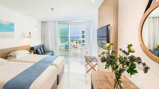 Et hotelværelse med to senge, en sofa, et skrivebord med blomster, stort spejl og en balkon med udsigt over havet. (AI-genereret tekst)