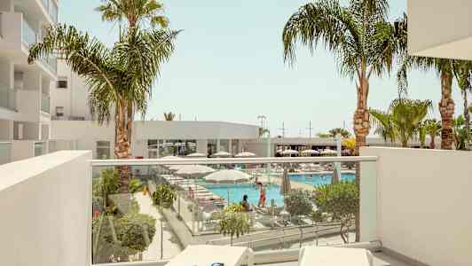 Udsigt fra en altan mod en pool omgivet af liggestole og palmer. (AI-genereret tekst)