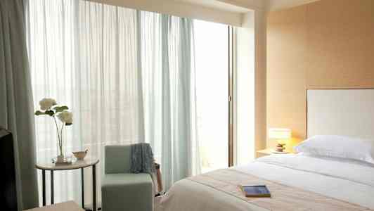 Et soveværelse med en redt seng, en stol, en bordlampe og et vindue med lyse gardiner. (AI-genereret tekst)