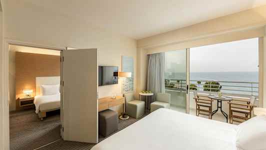 Et hotelværelse med en seng, siddepladser og et bord, samt en balkon med udsigt over havet. (AI-genereret tekst)