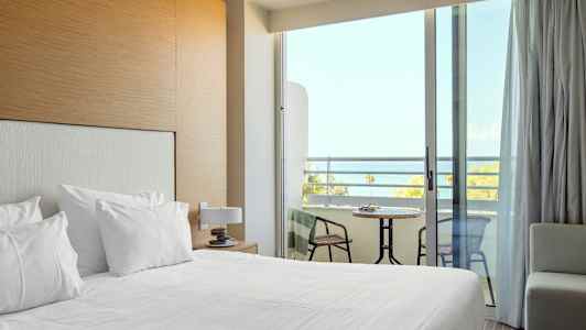 Et hotelværelse med redt seng og en balkon med udsigt over havet. (AI-genereret tekst)