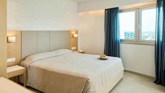 Et hotelværelse med en stor seng, to natborde, to lamper, et vindue med blå gardiner og en hat på sengen. (AI-genereret tekst)