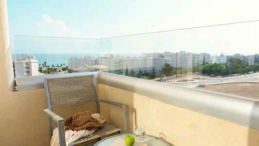 En altan med udsigt over havet og bygningerne, med en stol og et bord med glas, bog og et æble. (AI-genereret tekst)