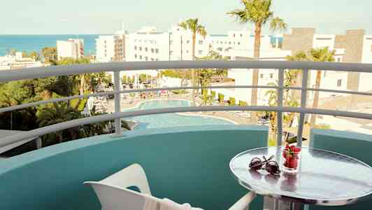 Udsigt fra en balkon med udsigt til en pool og omkringliggende bygninger; et lille bord har solbriller og et glas med jordbær. (AI-genereret tekst)