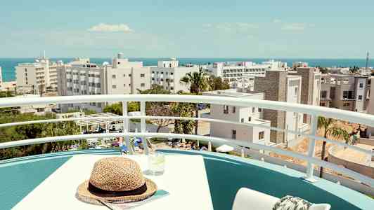 En balkon med bord, stol og udsigt over bygninger og havet i baggrunden. (AI-genereret tekst)