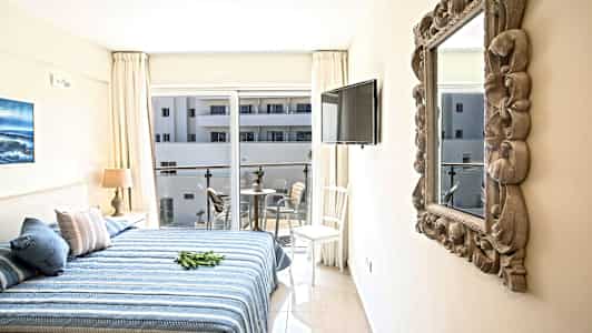 Et hotelværelse med en seng, bord og stole på en altan udenfor vinduet. (AI-genereret tekst)