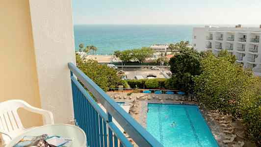 Udsigt fra en altan med bord og stol over en pool, grøntområder og havet. (AI-genereret tekst)