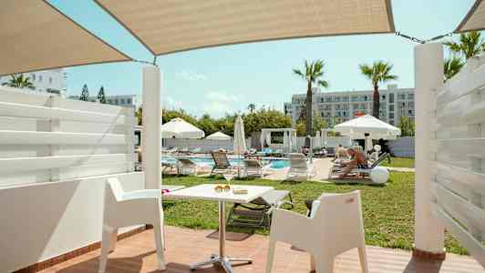 Udsigt fra en terrasse med hvide stole mod en pool omgivet af liggestole og parasoller. (AI-genereret tekst)