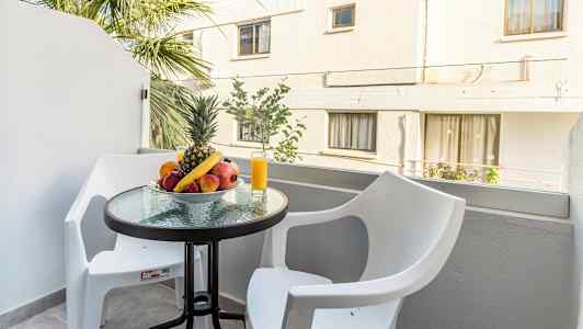 En altan med et bord, der har en skål med frugt og et glas med juice, omgivet af to hvide stole. (AI-genereret tekst)