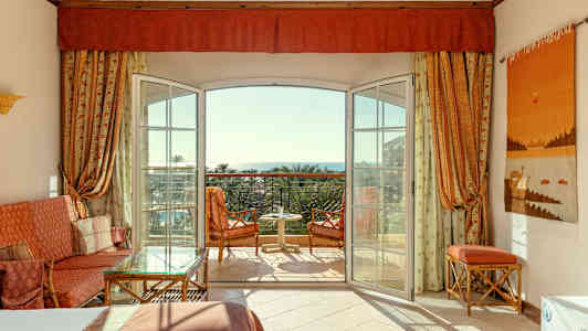 Et hotelværelse med åben altandør, siddepladser og udsigt mod havet. (AI-genereret tekst)