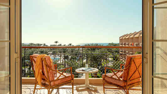 To stole og et bord står på en altan med udsigt over palmer og havet. (AI-genereret tekst)