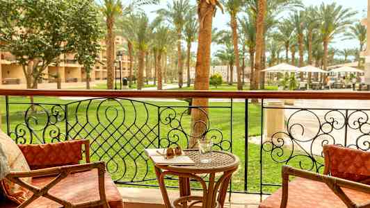 En balkon med stole, en bord med et glas og solbriller, og udsigt til en have med palmer. (AI-genereret tekst)