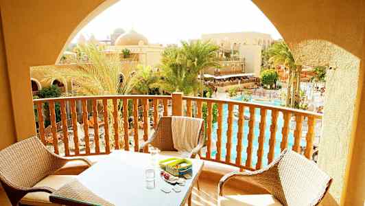 Altan med møbler med udsigt over palmer, pool og sandfarvede bygninger. (AI-genereret tekst)