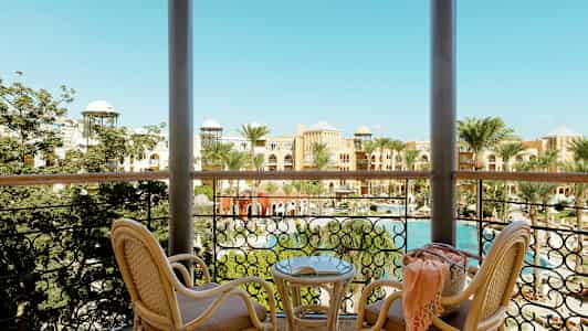 Udsigt fra en altan med to fletstole og et bord. Bag rækværket ses palmer, en swimmingpool og bygninger med farverige facader. (AI-genereret tekst)