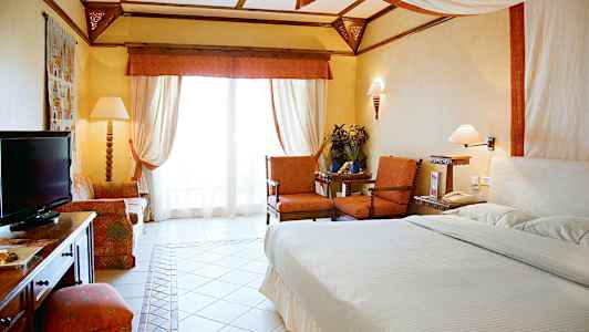 Et hotelværelse med en stor seng, to lænestole ved et lille bord, en sofa og et fjernsyn. Værelset er lyst og elegant indrettet. (AI-genereret tekst)