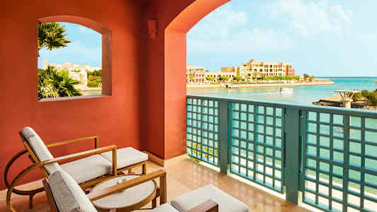 Udsigt fra en balkon med siddepladser, over en kystlinje og bygninger. (AI-genereret tekst)